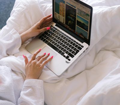 A woman is sitting on a bed with a laptop on her lap. She is wearing a white sweater and the bedding is also white. We can only see her hands, sweater, laptop, some of the bedding.