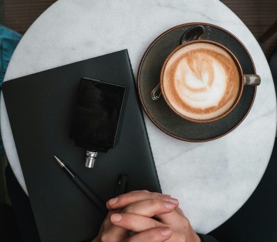 Image of a persons hands interlocking, resting on a black book. The book sits on a table with a latte that is in a black cup.
